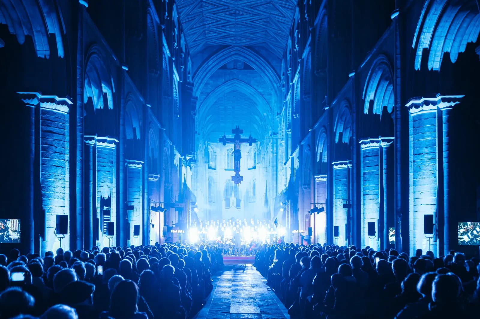 A large crowd of people sits facing a brightly lit stage in a grand, blue-lit cathedral with a large cross hanging above.
